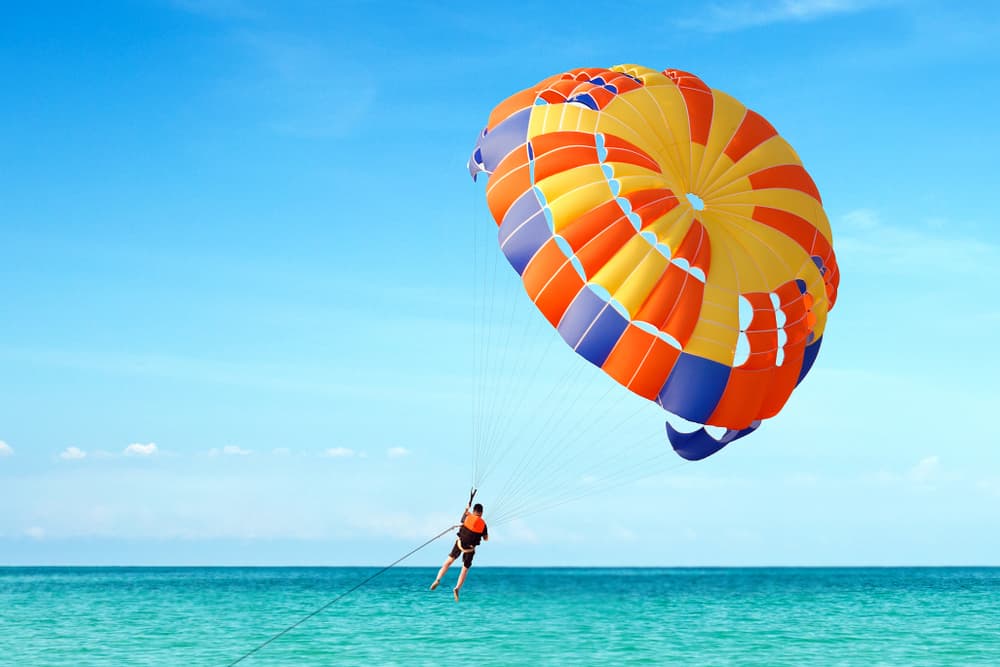 A person parasailing over the ocean, attached to a colorful parachute with a clear blue sky in the background.