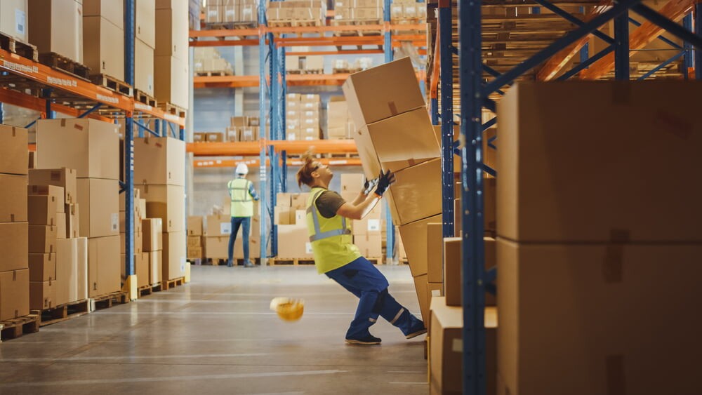 warehouse worker in yellow safety vest struggles under stack of boxes actively falling on top of him