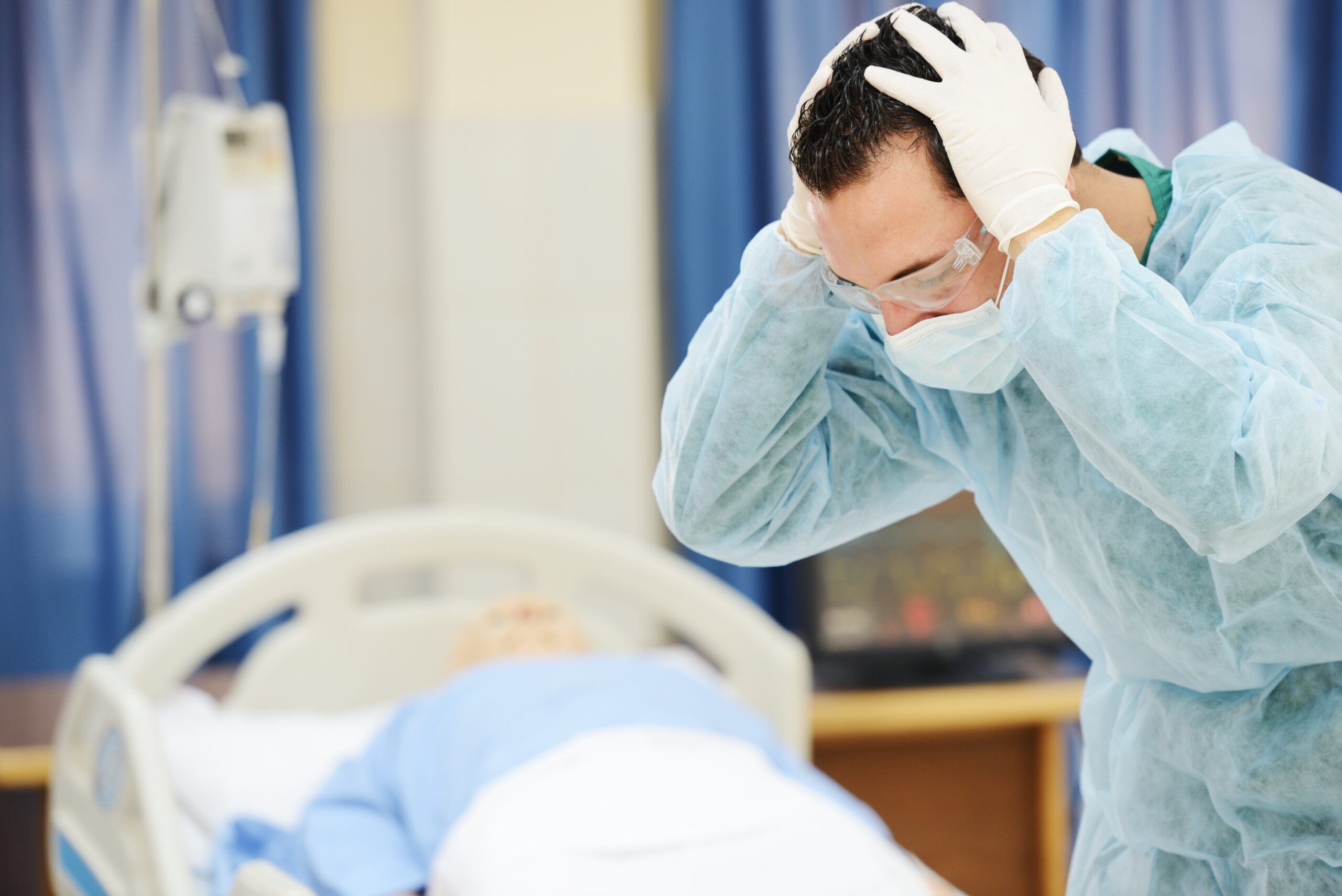 doctor with his head in his hands standing in front of a sleeping man on a hospital bed