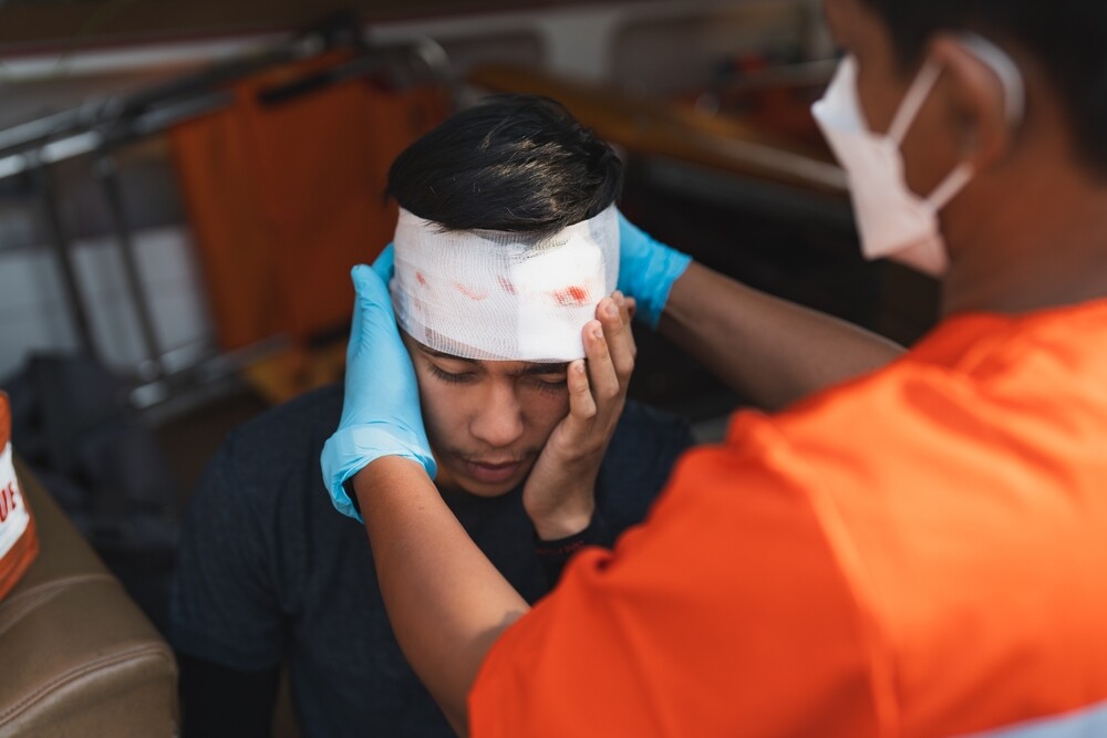 injured man holding his hand to his head while another man dresses patients head wounds