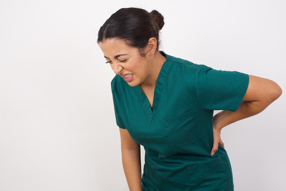woman in green scrubs holding her lower back and wincing in pain
