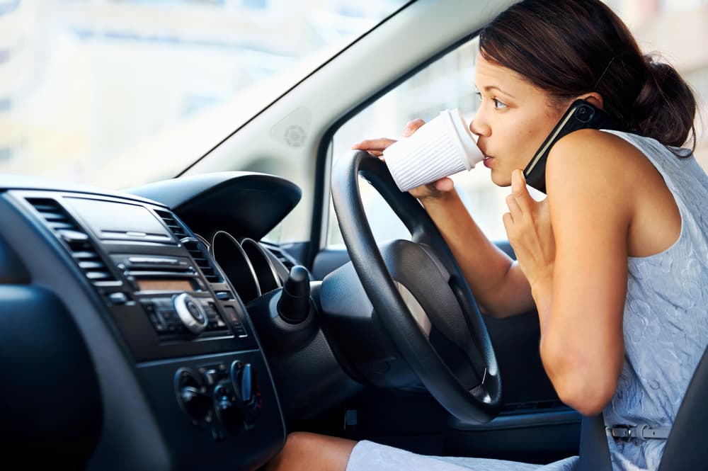 A woman is driving while distracted, drinking from a white cup and talking on a mobile phone held between her shoulder and ear. The car's dashboard is visible.