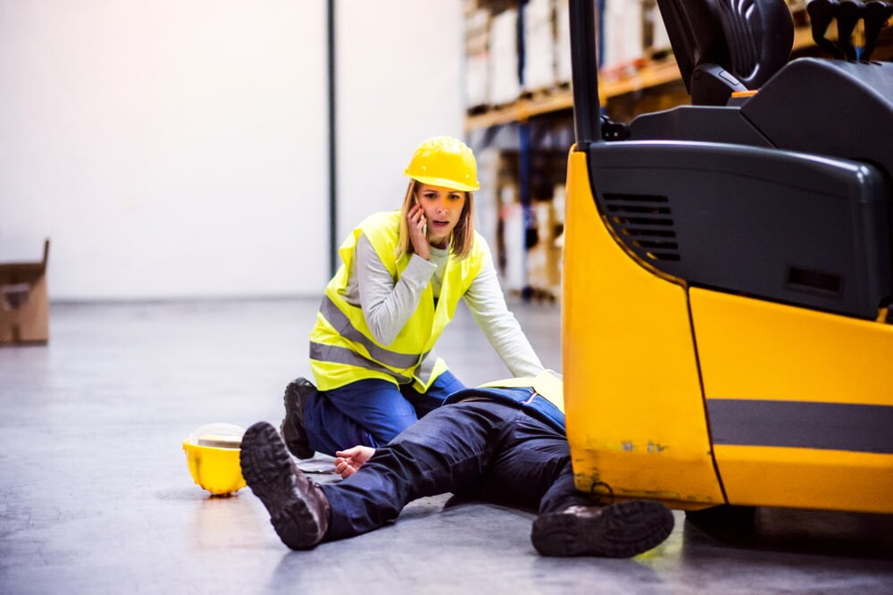 man laying down with his leg pinned under a forklift and a female coworker next to him calling someone while looking panicked